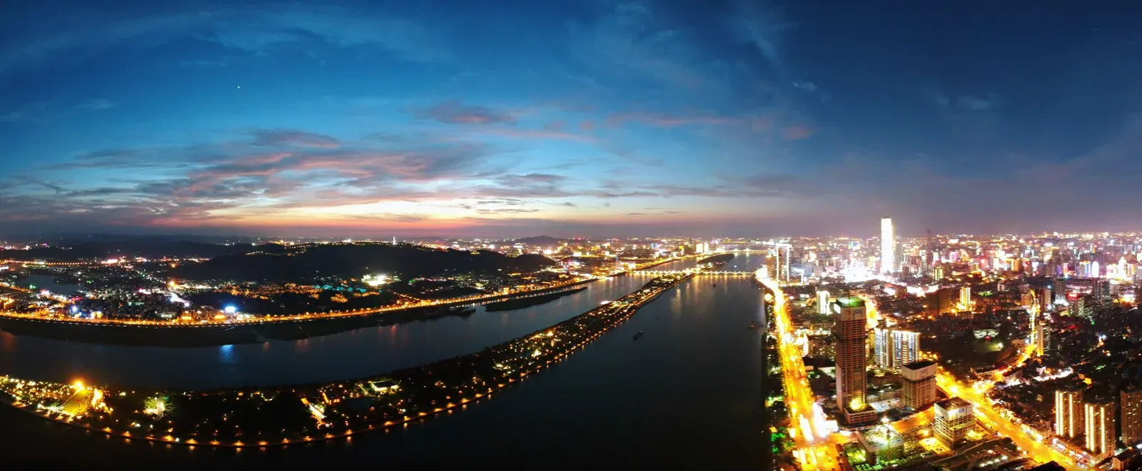 Panorama of a river city at dusk, both banks lit.