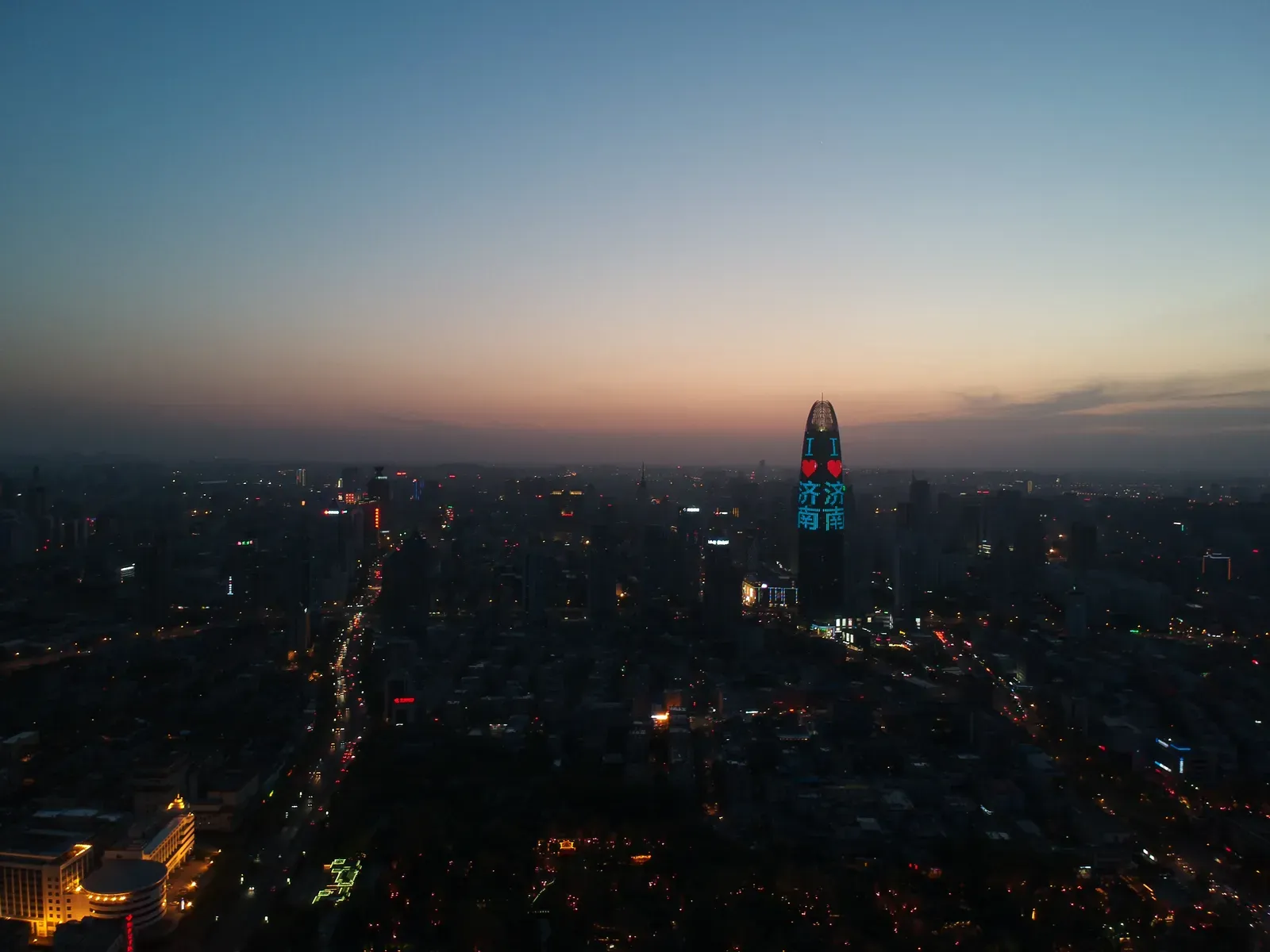 City at dusk from the air, one tower lit blue.